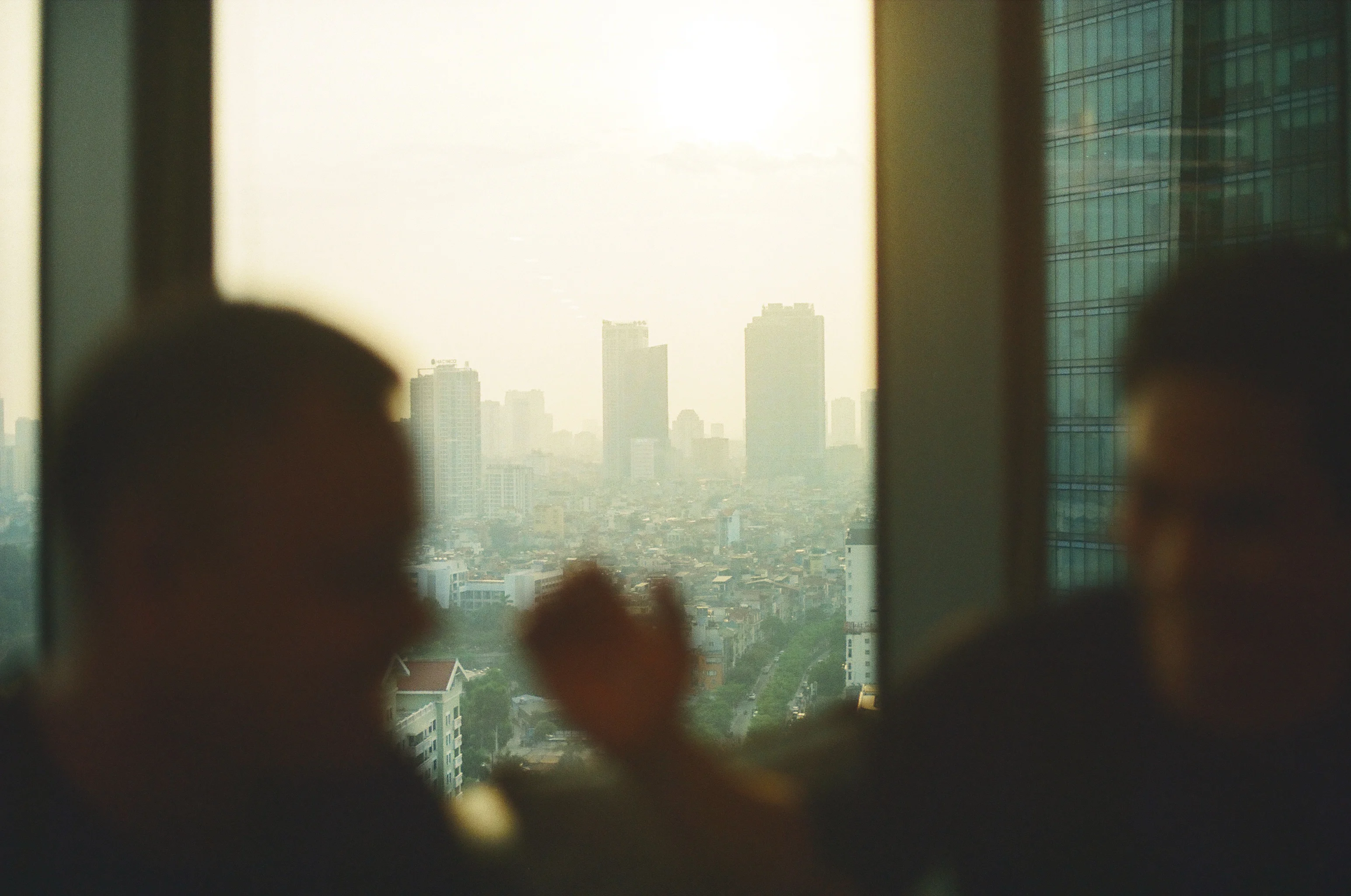 Two blurred silhouettes of people in front of a large window overlooking a hazy urban skyline with high-rise buildings.