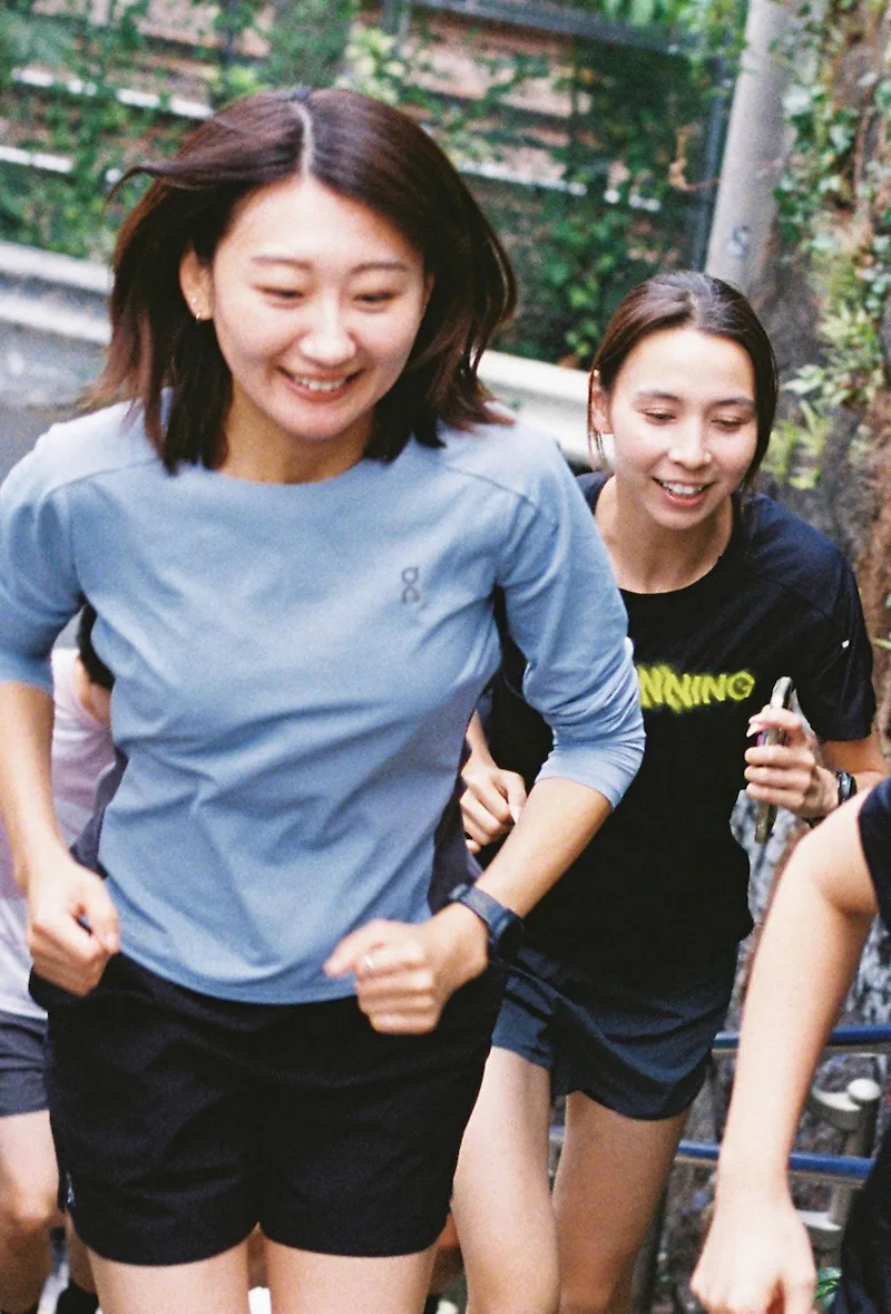 Two women in activewear run outdoors, smiling, surrounded by greenery and other runners.