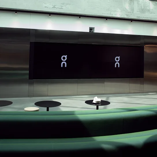 Modern interior with curved green bench seating, round black tables, a large black wall-mounted screen showing two identical white stylized symbols, and a small white object on one table.