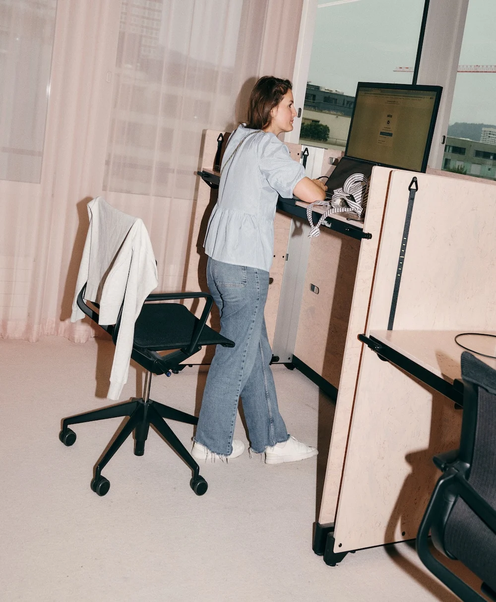 A woman stands at a wooden standing desk in an office cubicle using a computer, with a jacket draped over a nearby office chair.