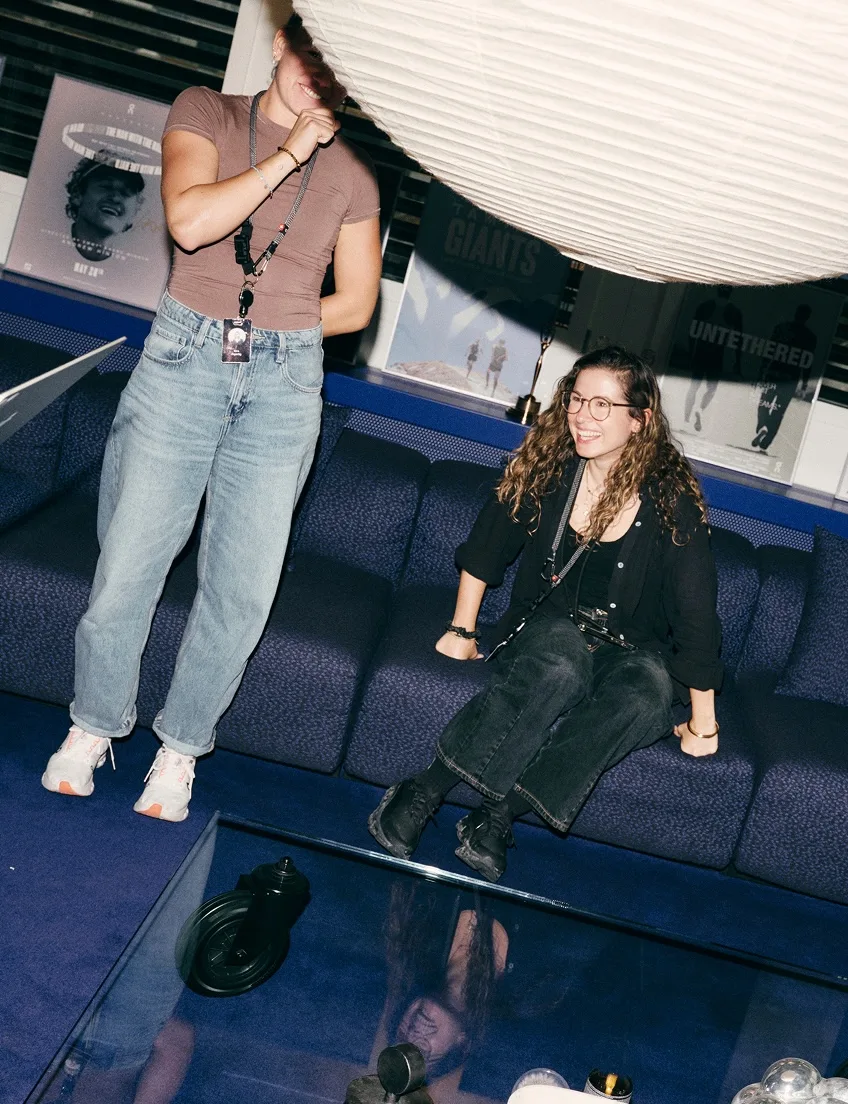 Two women in casual clothes talk and laugh in a lounge with a large hanging light and film posters in the background.