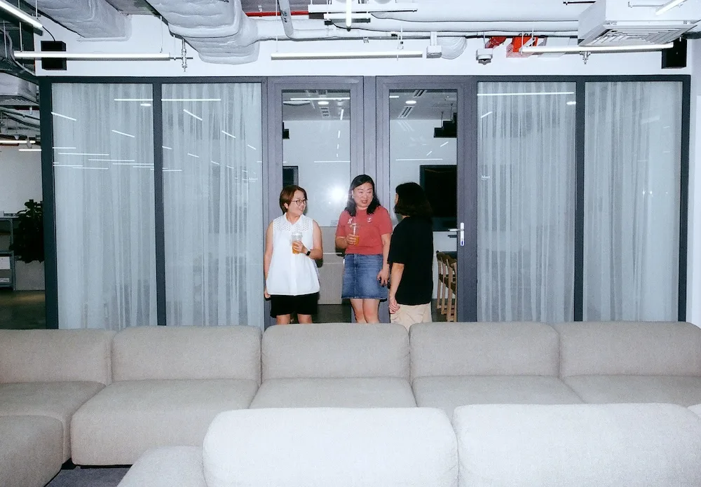 Three people stand talking and holding drinks in front of glass doors with curtains in an office lounge, with beige sofas in the foreground.
