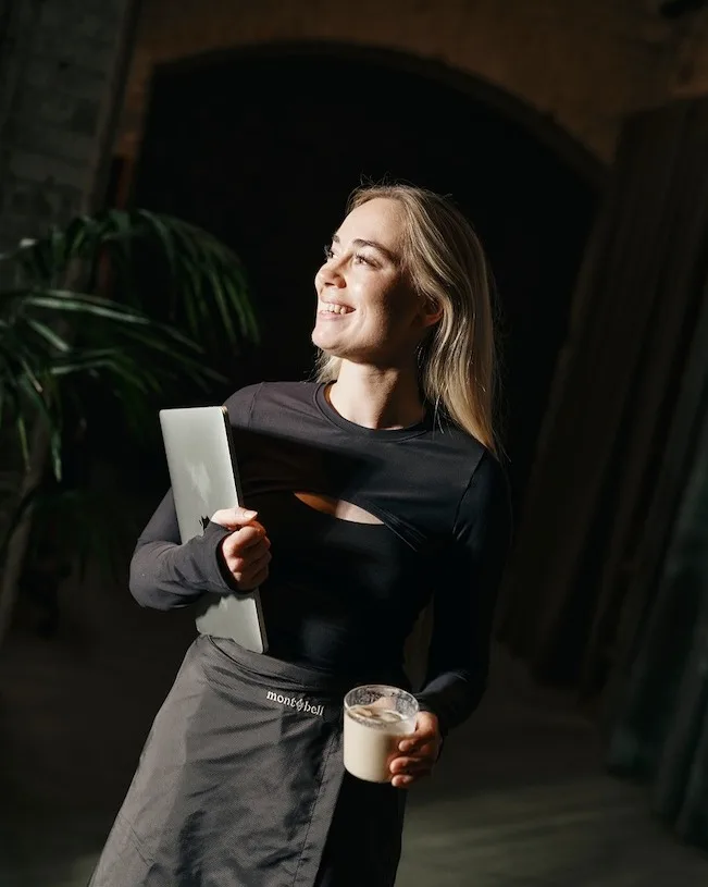 A smiling woman with long blonde hair stands indoors holding a closed laptop under her arm and a glass of a light-colored drink, wearing a dark long-sleeve top and a gray apron while looking up and to the side.