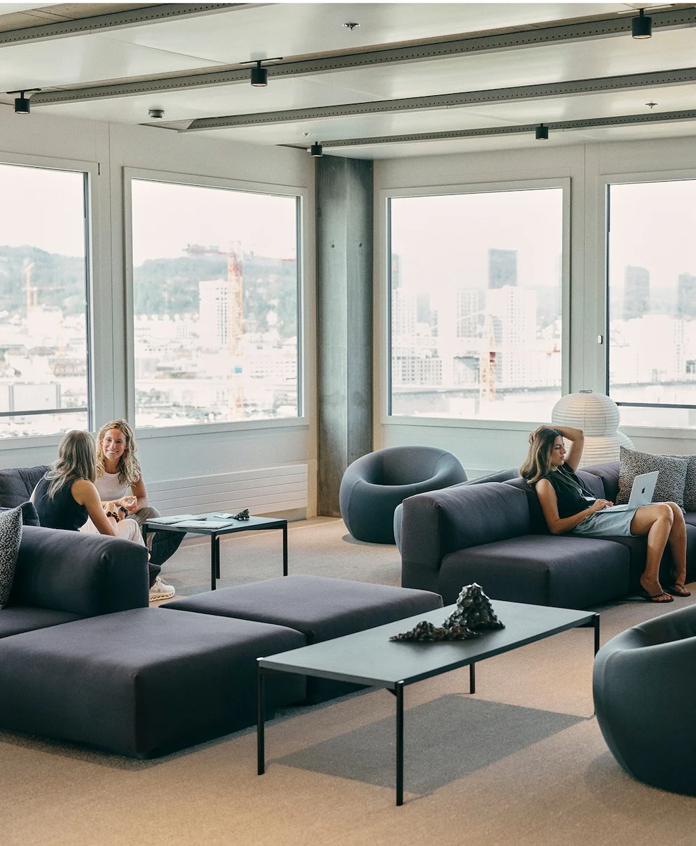 Three women in a modern lounge with large windows and dark modular sofas, two talking at a low table and one reclining on a sofa using a laptop.