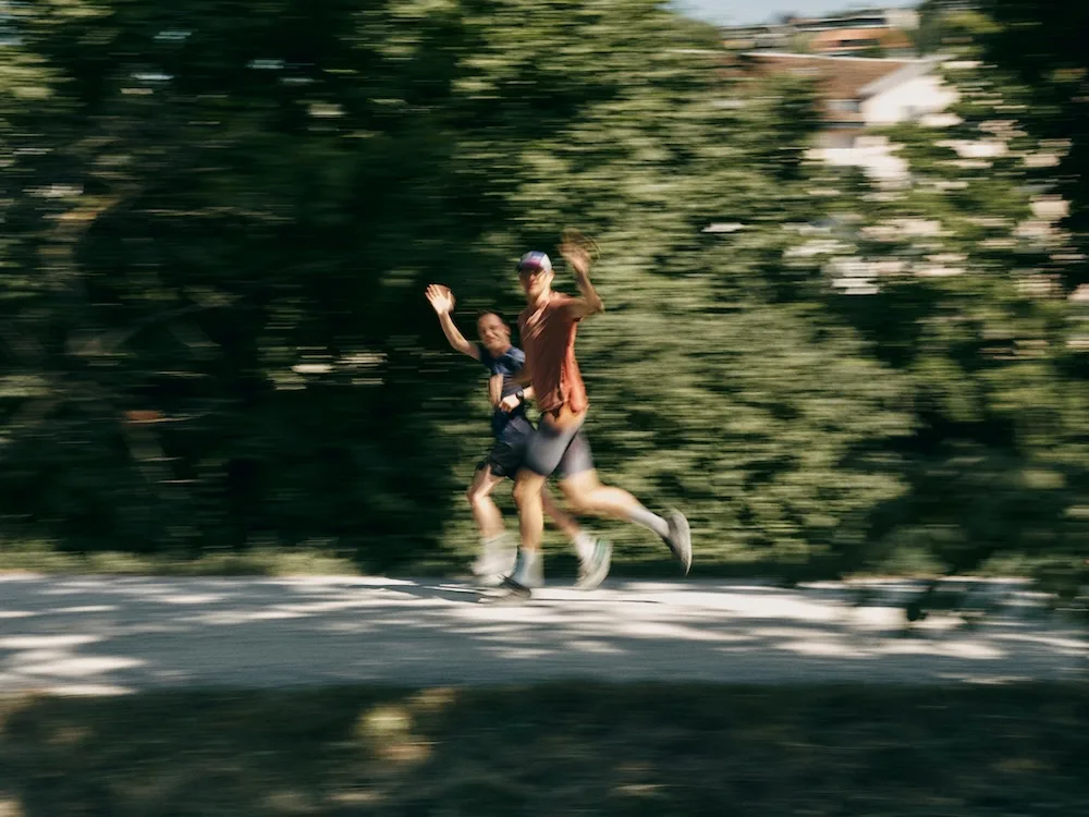 Two runners wave and smile while jogging on a shaded path, captured in motion blur against a backdrop of trees.