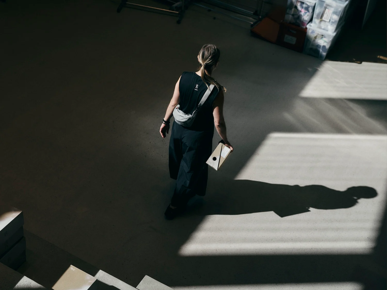 Woman in a black outfit walks across a sunlit room carrying a small cardboard box, casting a strong shadow on the floor.

