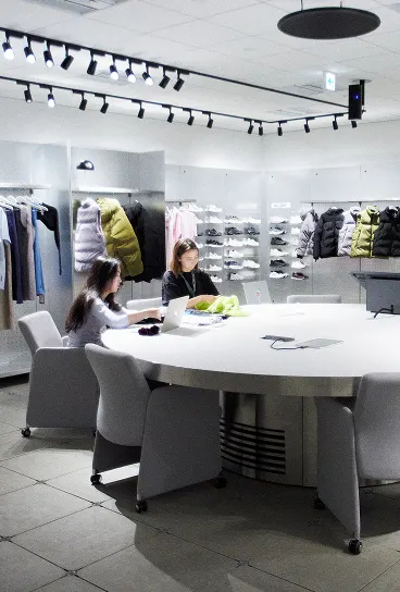 Two women sit at a large round table with laptops in a brightly lit clothing and shoe showroom where jackets and sneakers are displayed on wall-mounted shelves.