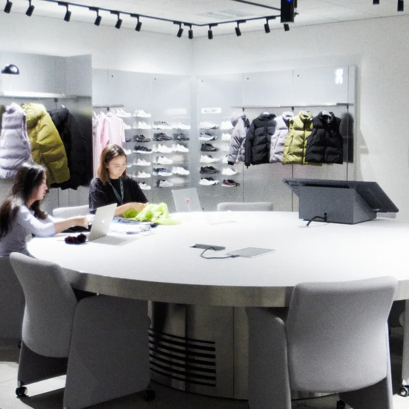 A bright room with a large round white table and chairs in the foreground and a wall display of shoes and four puffer jackets hanging above a counter in the background.