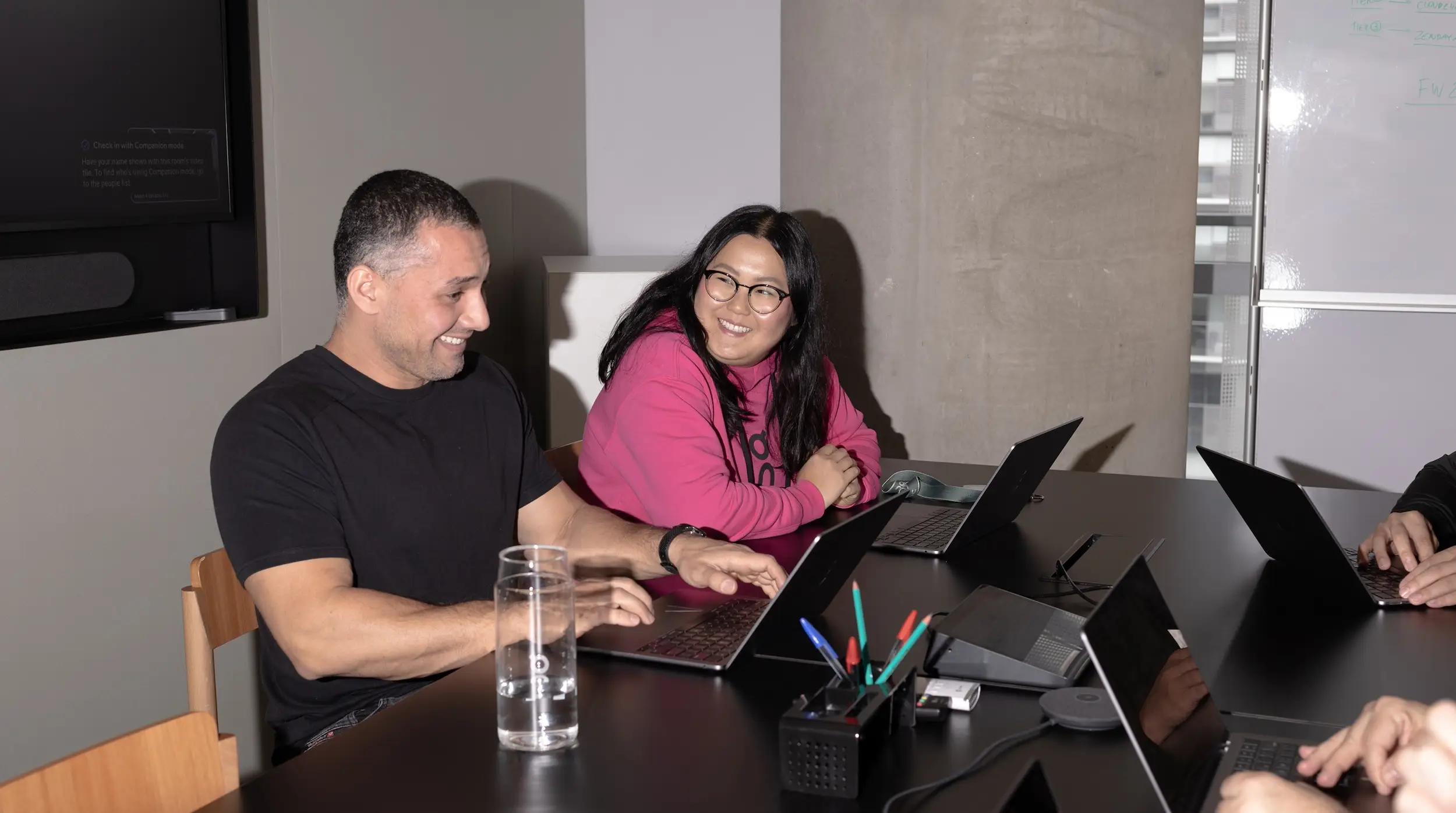 Two colleagues, a man in a black t-shirt and a woman in a pink hoodie with glasses, sit at a conference table smiling while using laptops. 