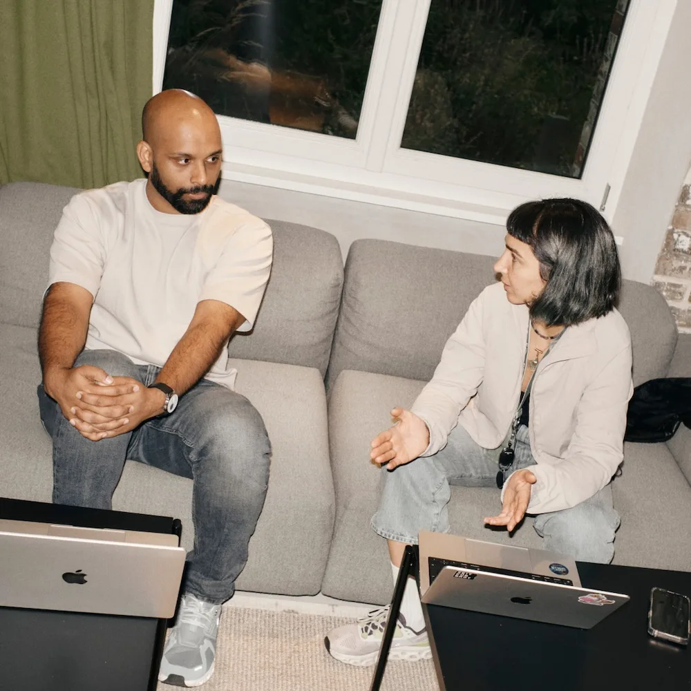 Two people talk on a grey couch with laptops on black tables, engaged in discussion near a window.