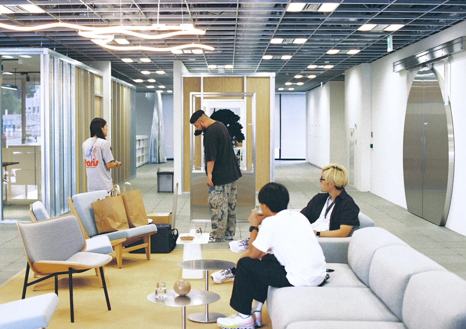 Four people relax and chat in a lounge area with light furniture and shopping bags, under modern ceiling lighting.