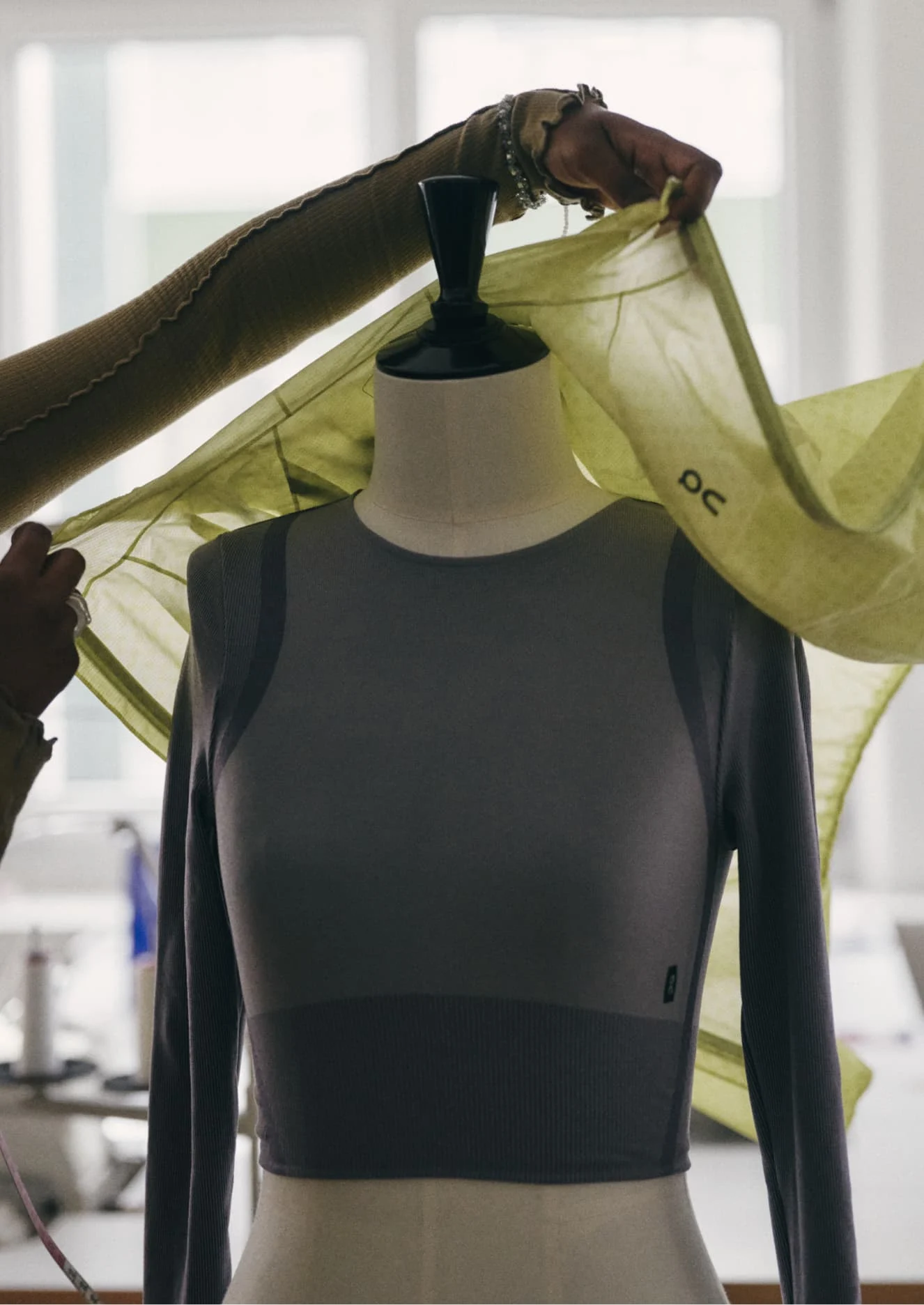 Fashion designer drapes a light green On-branded fabric over a fitted grey top on a dress form in a bright studio.