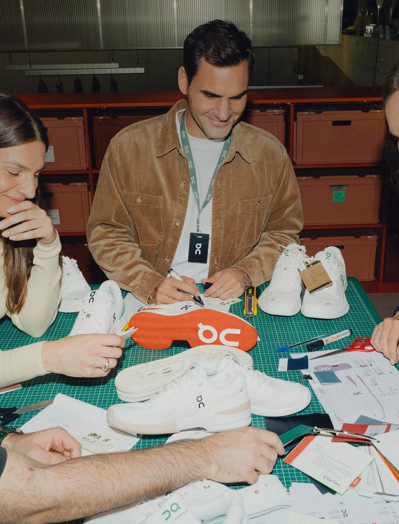 A man in a brown corduroy jacket and a lanyard sits at a worktable with colleagues, examining and marking white athletic shoes and orange shoe soles.