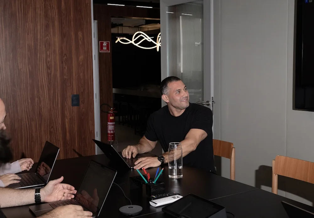 A man in a black T-shirt sits at a conference table with a laptop and a glass of water, smiling and looking to his right while colleagues work on laptops.