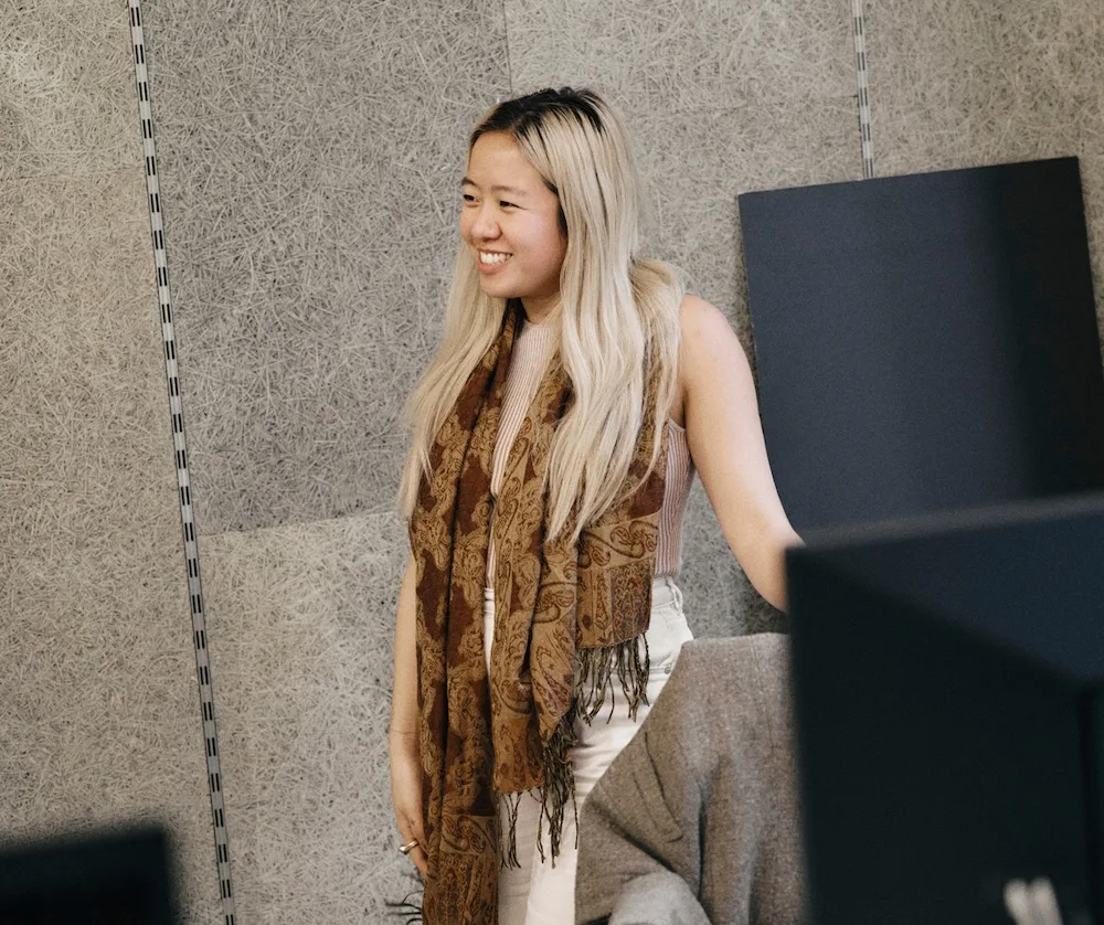 A smiling woman with long blonde hair stands in a workspace wearing a patterned brown scarf, sleeveless top, and light-colored pants.