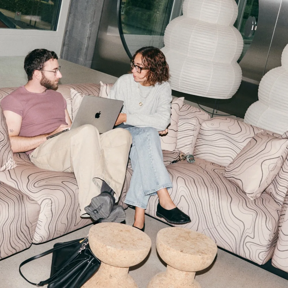 Man and woman sit on a patterned couch with a laptop, talking in a modern lounge with sculptural white lamps.