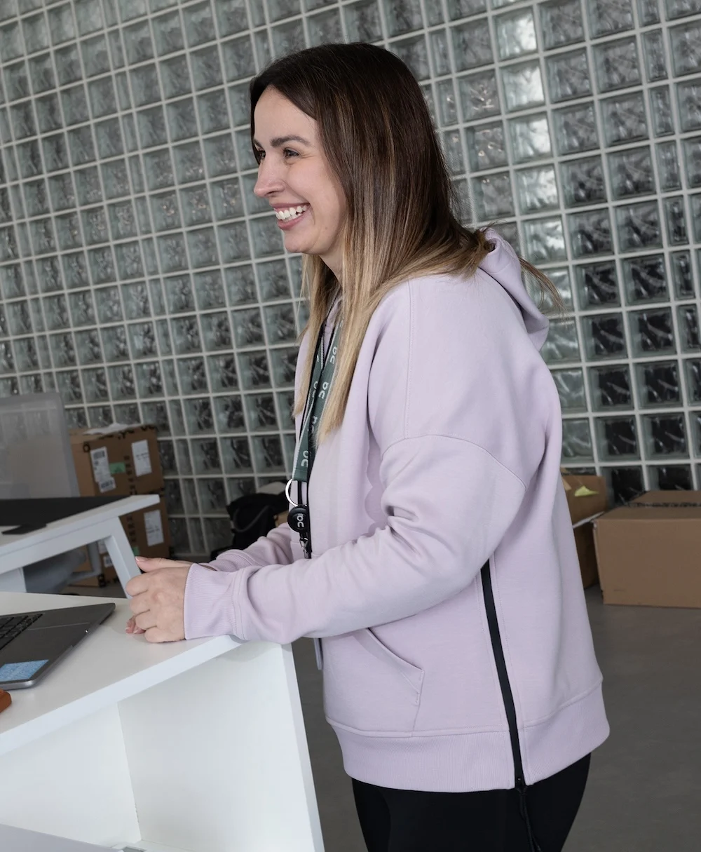 A smiling woman in a light purple hoodie and lanyard leans on a white counter with a laptop in front of a glass-block wall.