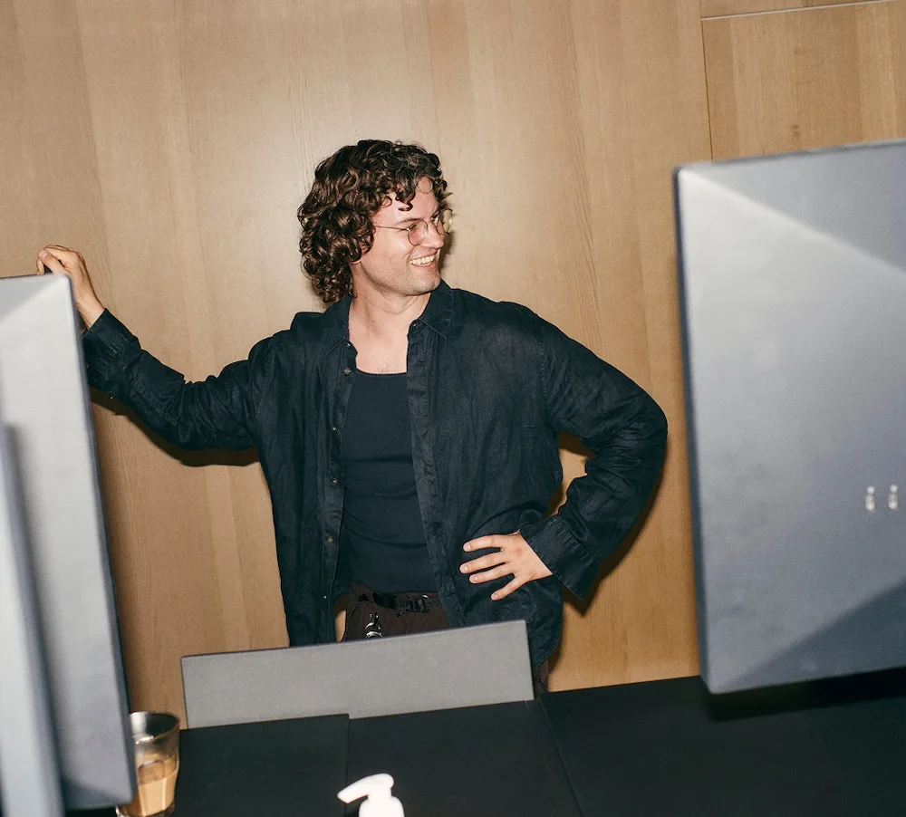 A smiling person with curly hair and glasses stands between two large computer monitors in front of a wood-paneled wall with one hand on their hip.