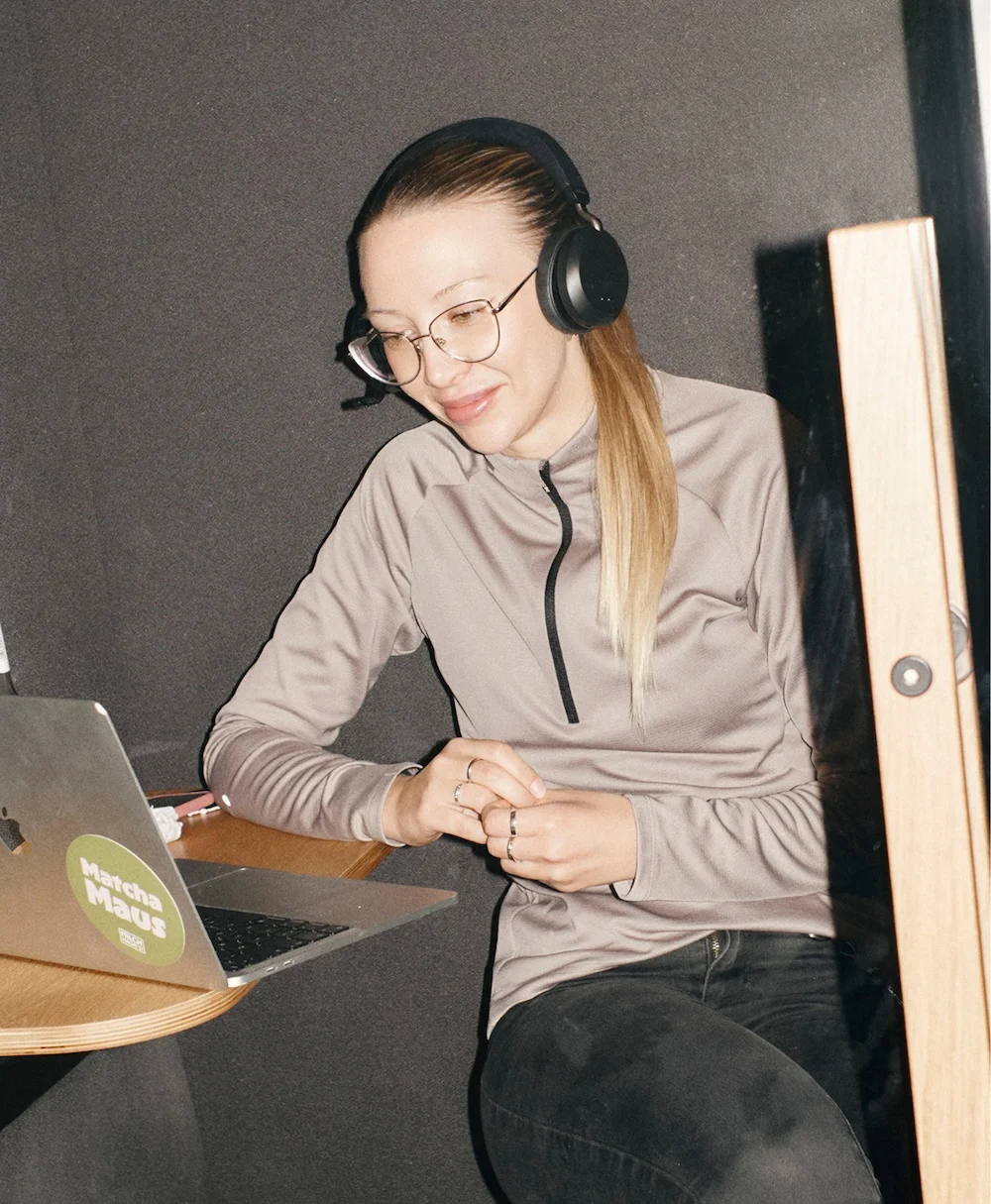 A person wearing glasses and over-ear headphones sits in a small booth at a wooden desk, looking at a laptop with a green "Matcha Maus" sticker.