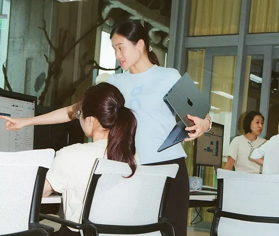 A standing woman holding a laptop points at a computer screen while a seated woman watches in an office.