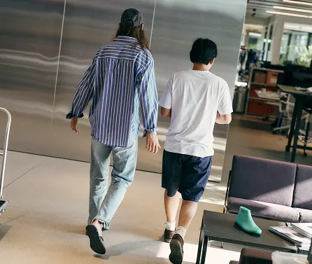 Two people are walking away down an indoor space, seen from behind—one wearing a blue striped shirt, jeans and a baseball cap, the other wearing a white T-shirt and dark shorts.