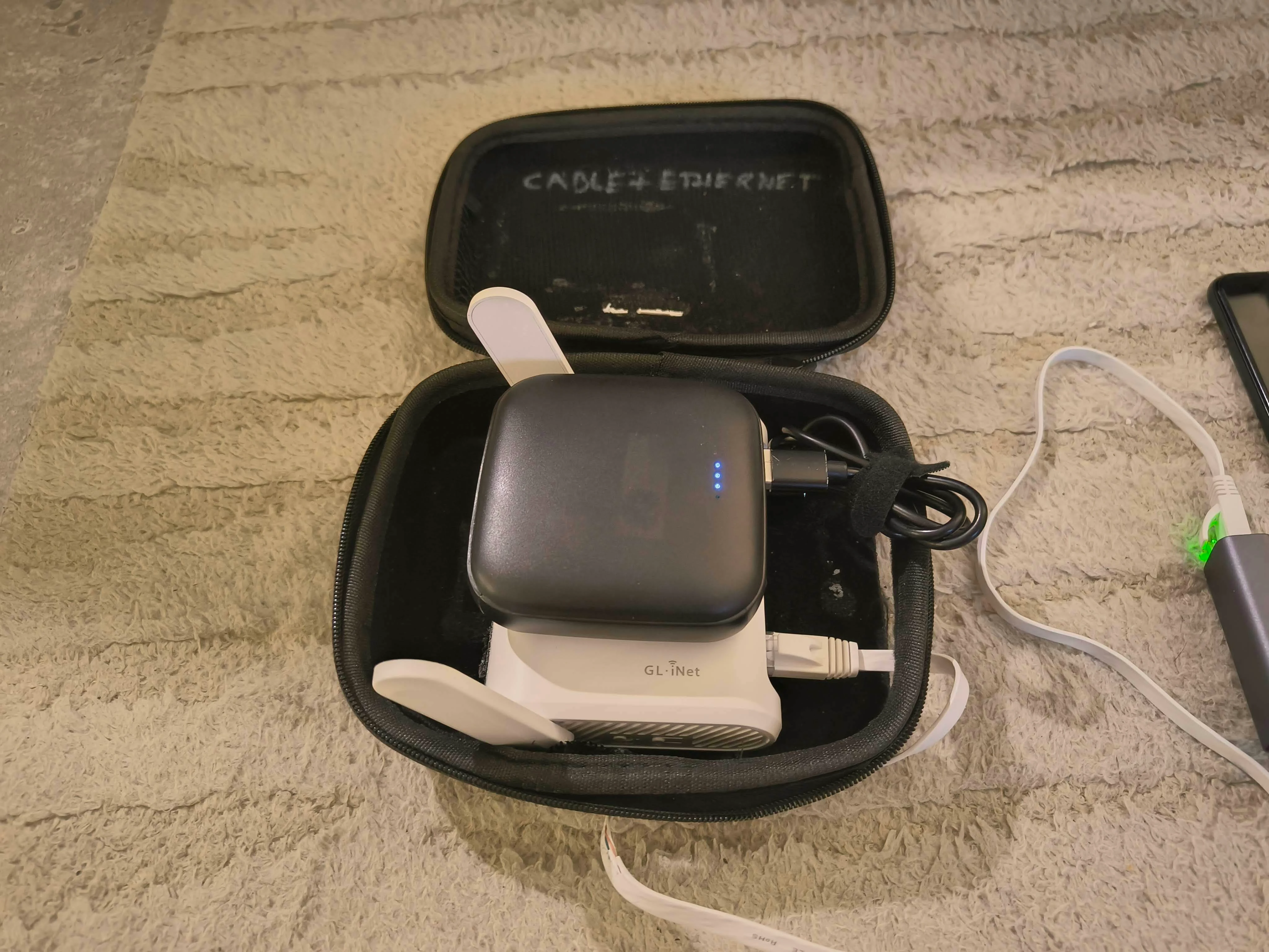Photo of the carry case open showing GL.iNet router and power bank inside, labeled CABLE + ETHERNET