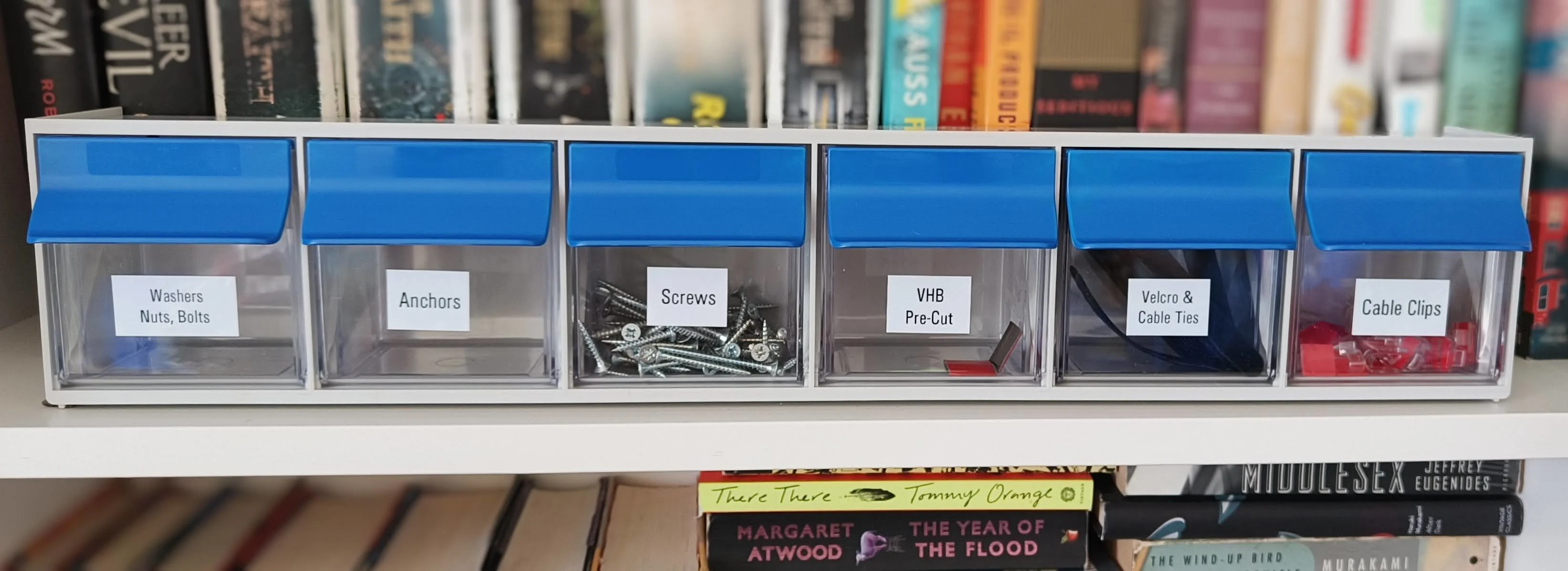 A row of small picking bins mounted over a bookshelf: washers, anchors, screws, VHB, velcro, cable clips.