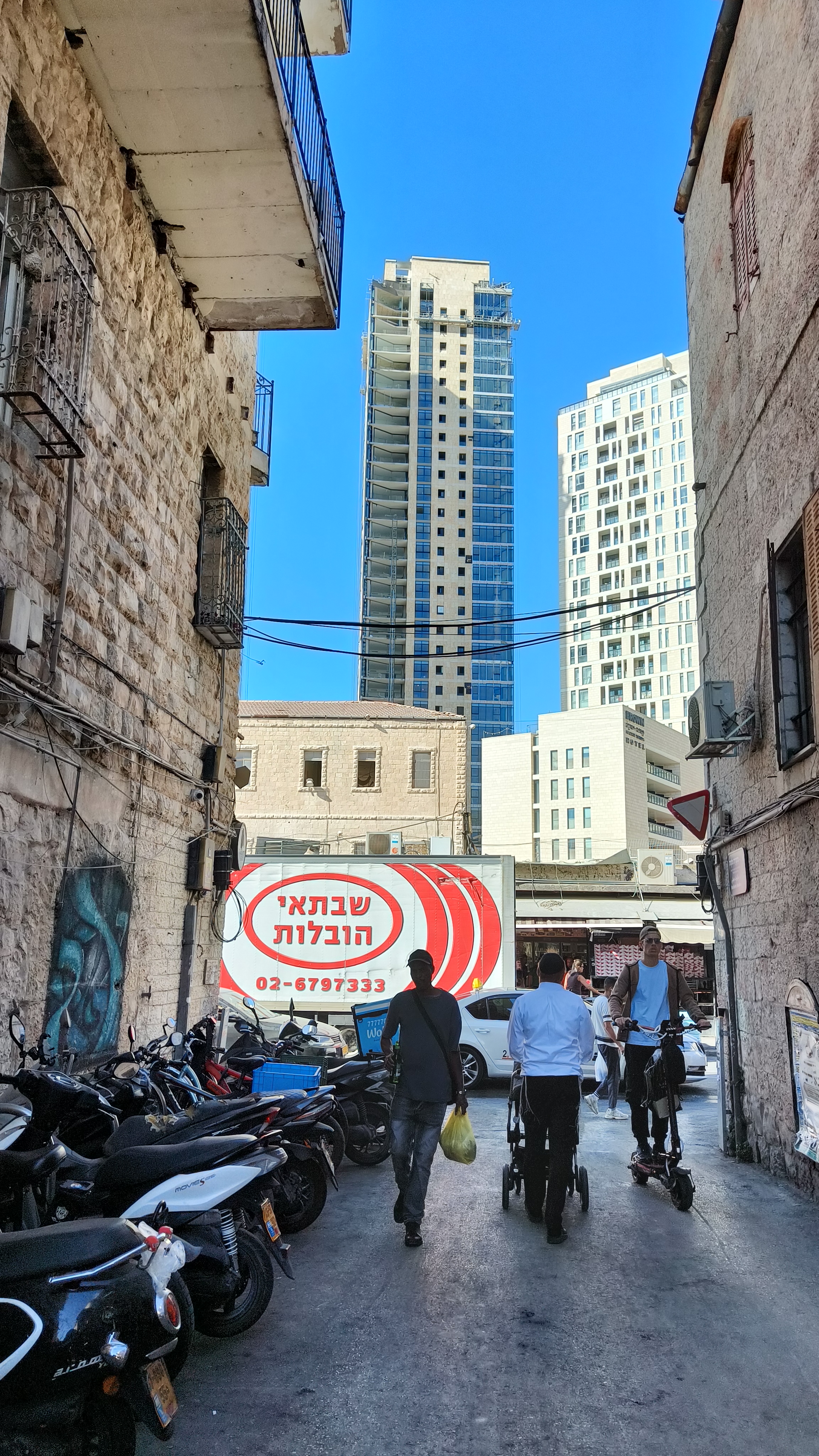 Pedestrians walk from an alleyway in meandering Nahlaot towards the shuk with the high rises of the Kiach compex towering in the background. Photo: author.