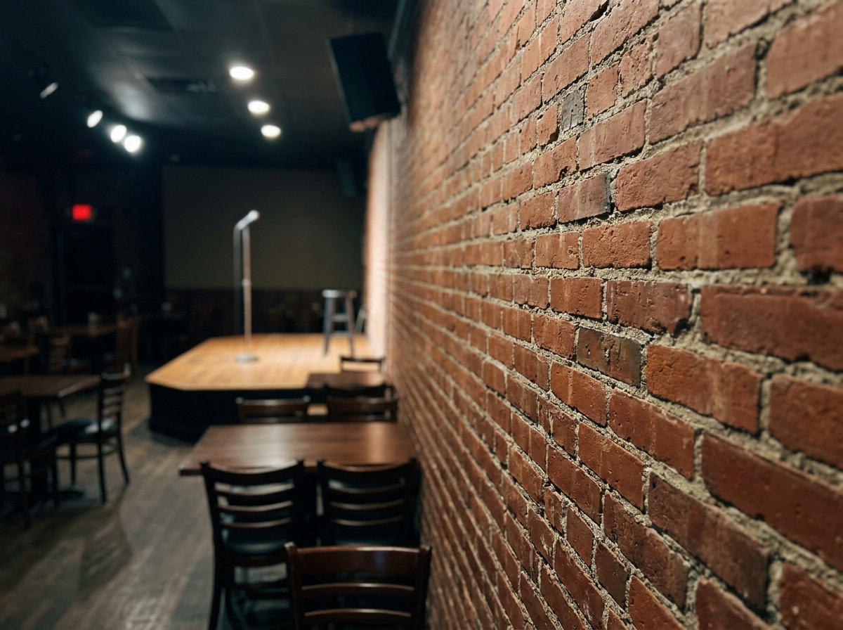 A brick wall inside a comedy club, with the empty stage slightly out of focus in the background.