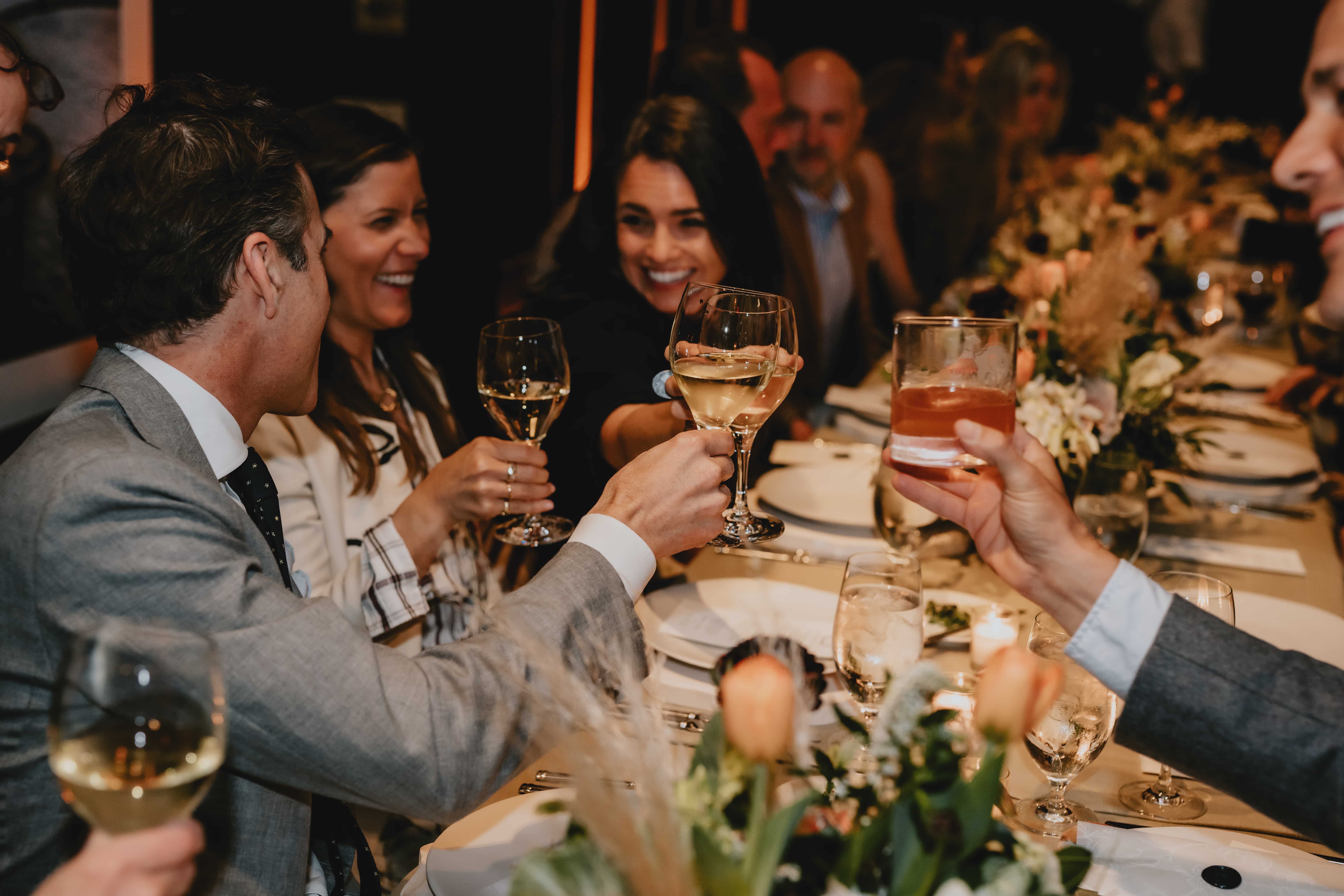 A group of people seated at a long, elegantly decorated table raise their glasses for a toast.