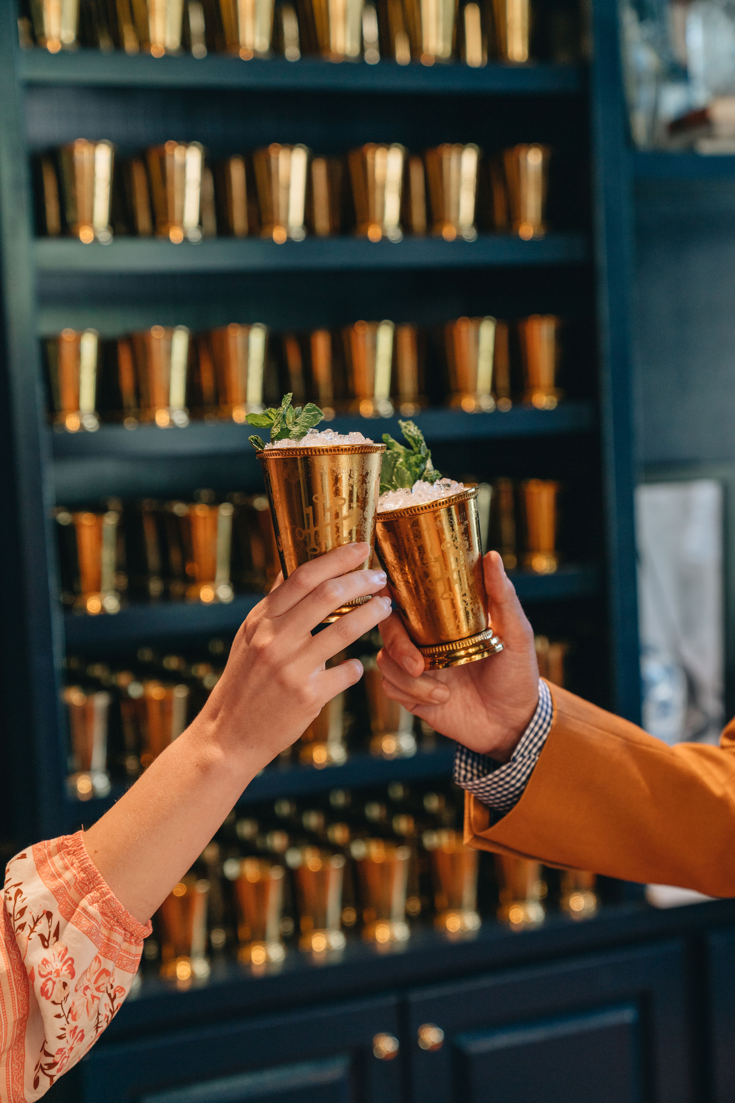 Two hands toasting with gold cups filled with ice and mint leaves, against a backdrop of similar cups on shelves.