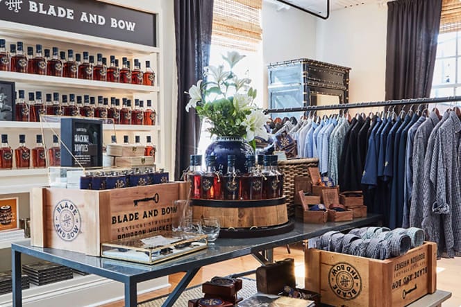 A stylish boutique store interior featuring a display of Blade and Bow Kentucky Straight Bourbon Whiskey bottles arranged on shelves and wooden crates.