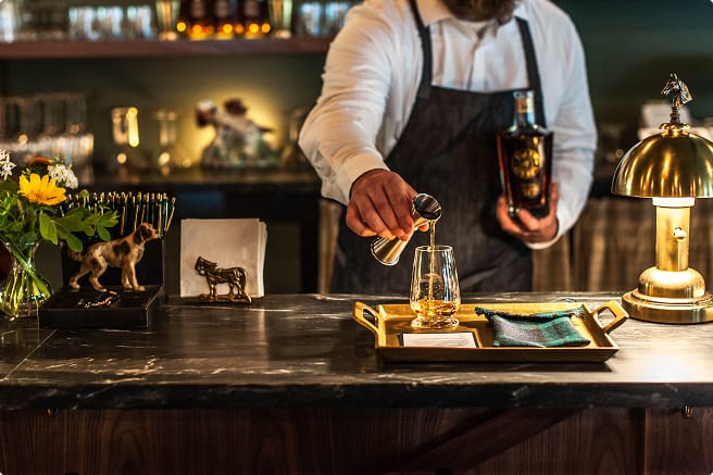 A bartender in a white shirt and dark apron pours whiskey from a jigger into a glass on a tray.