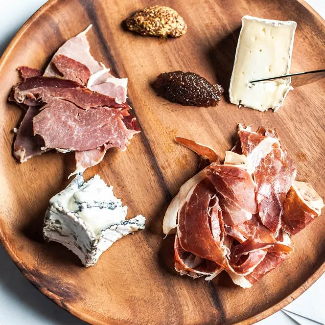 A wooden platter featuring an assortment of gourmet foods, including slices of cured meats, two types of soft cheeses, and dollops of mustard and fig jam.