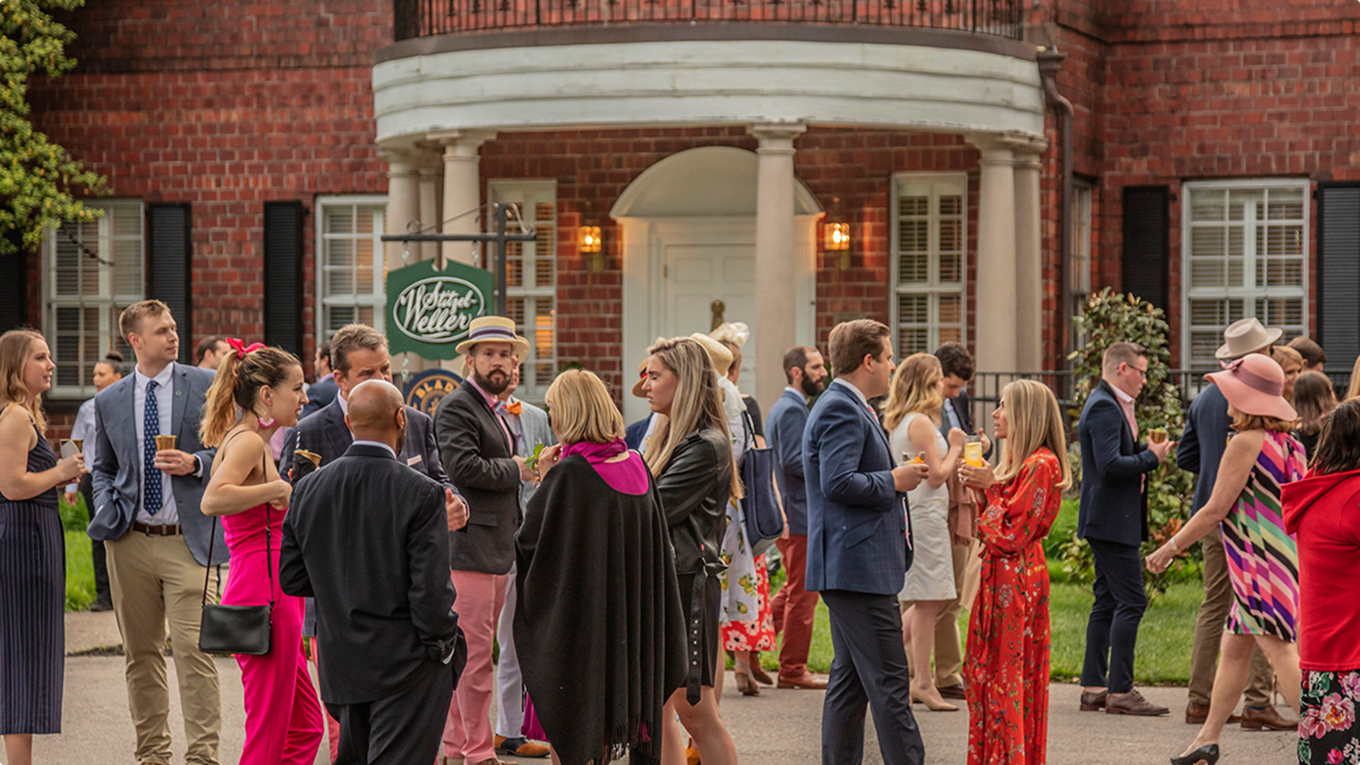 A lively outdoor gathering in front of a grand brick building with white columns. Guests are dressed in colorful and formal attire, mingling and holding drinks in a garden setting.