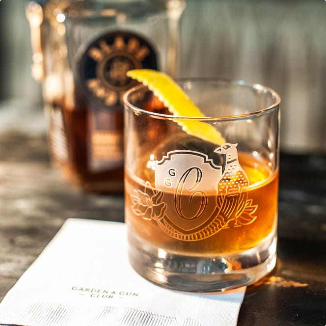 A glass of whiskey with a lemon twist sits on a table next to a branded napkin that reads "Garden & Gun Club."