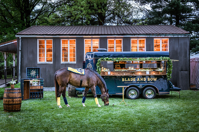A horse grazes on a lush green lawn in front of a stylish trailer bar labeled "Blade and Bow," which is decorated with greenery and lights.