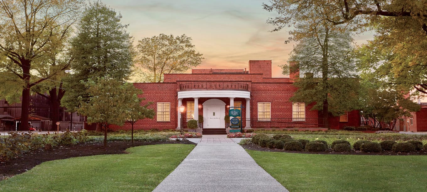 Historic Stitzel-Weller distillery entrance with red brick facade, white columns, and surrounding greenery at sunset.