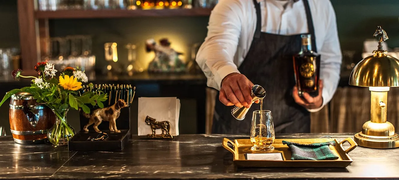 Bartender pours whiskey into a glass on a tray. Flowers, horse figurine, and lamp decorate the bar counter.