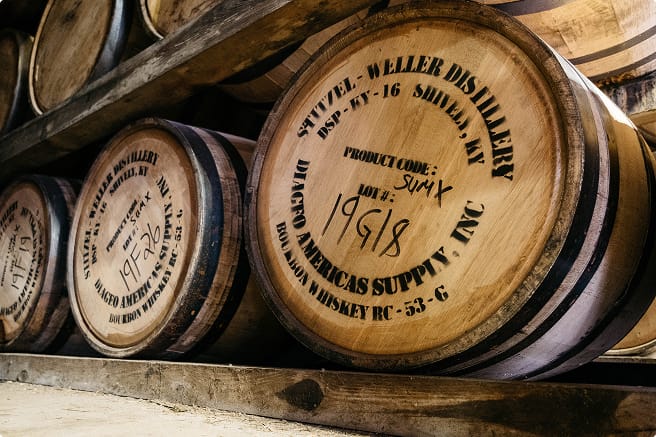 A row of wooden whiskey barrels stored in a distillery. The barrels are marked with inscriptions including "Stitzel-Weller Distillery," "Shively, KY," and "Diageo Americas Supply Inc."