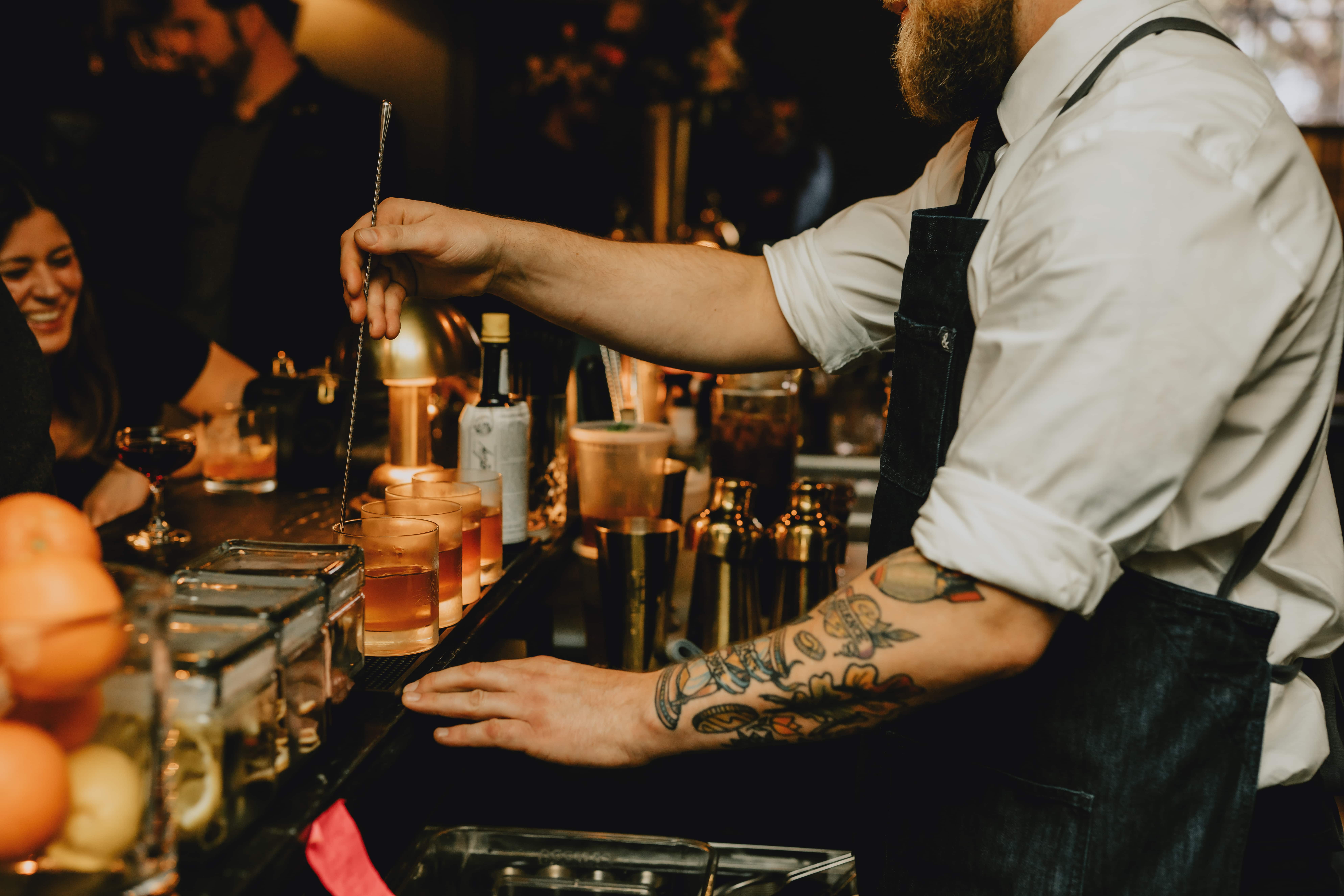 A bartender wearing a white shirt and dark apron, with tattooed arms, is stirring cocktails at a bar.
