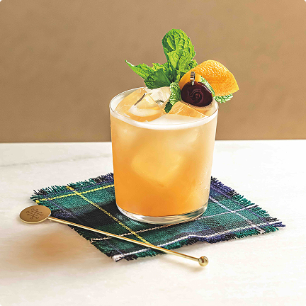 A glass filled with an orange-colored iced beverage sits on a green-and-blue tartan-patterned cloth.