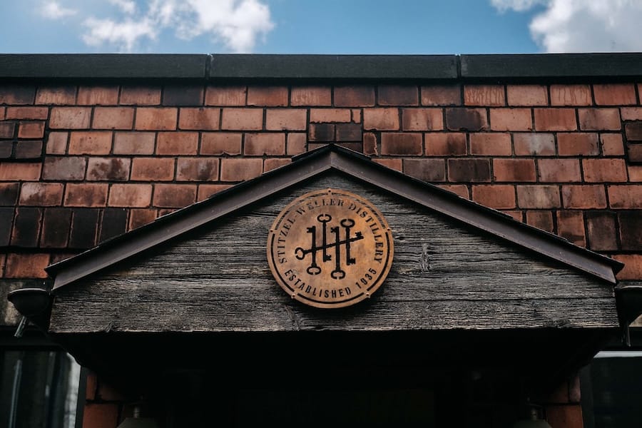 Weathered wooden gable with Stitzel-Weller Distillery emblem, established 1935. Red brick wall and cloudy blue sky in the background.