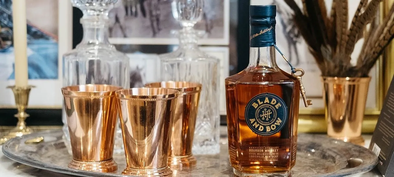 A bottle of Blade and Bow whiskey is placed on a silver tray alongside three copper cups. Behind the tray, there are crystal decanters and framed photographs.