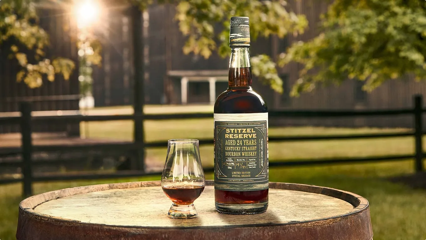 A bottle of Stitzel Reserve aged 24 years Kentucky Straight Bourbon Whiskey sits on a wooden barrel outdoors, accompanied by a filled whiskey glass.