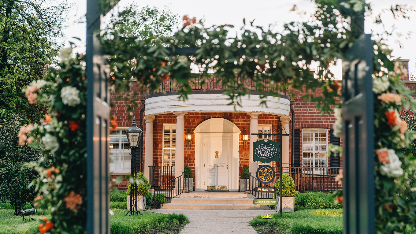 A view through a flower-decorated archway looking toward a brick building with white columns framing the entrance.