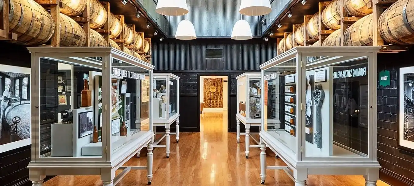 An interior view of the Stitzel-Weller Distillery showcasing a gallery with display cases. The room is lined with wooden barrels on shelves above the exhibits.