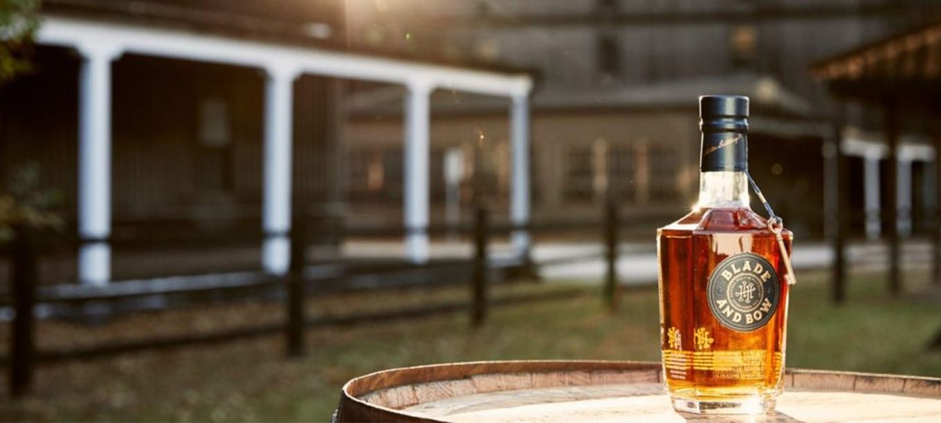 A bottle of Blade and Bow whiskey sits atop a wooden barrel outdoors, with the historic Stitzel-Weller Distillery in the background.
