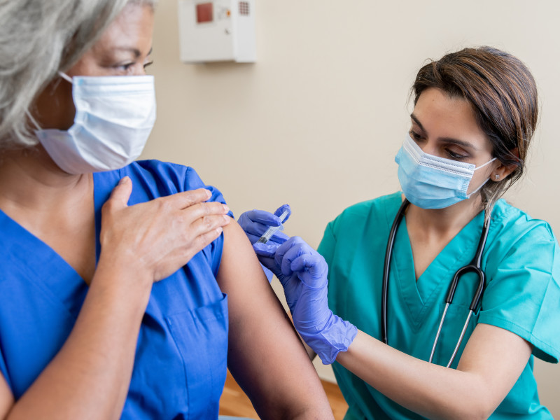 Young masked female nurse in teal scrubs gives a shot to an older female patient wearing a mask and a blue v-neck shirt.