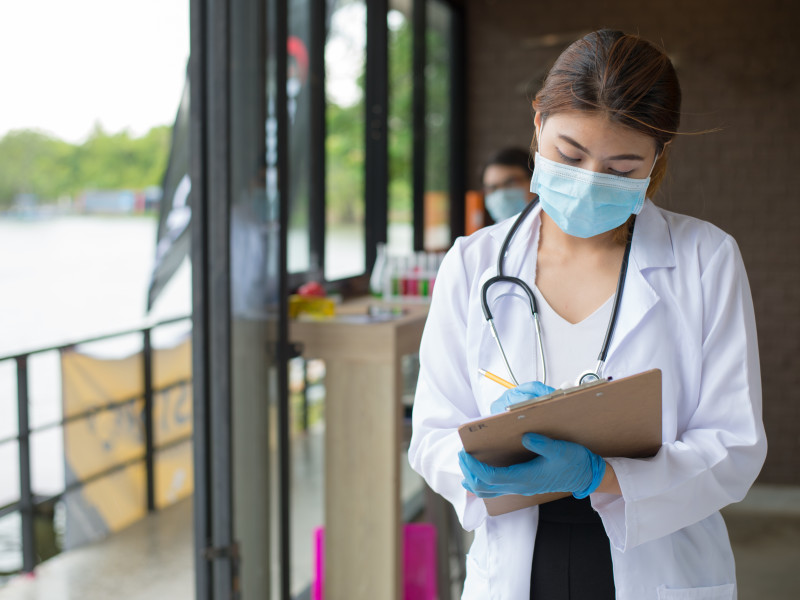 Young Asian-American female nurse wearing mask, white lab coat, and stethoscope taking notes on a clipboard in the middle of a hallway.